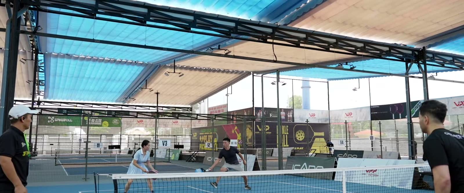 Hai người chơi pickleball trên sân có mái che, biển quảng cáo chạy quanh sân