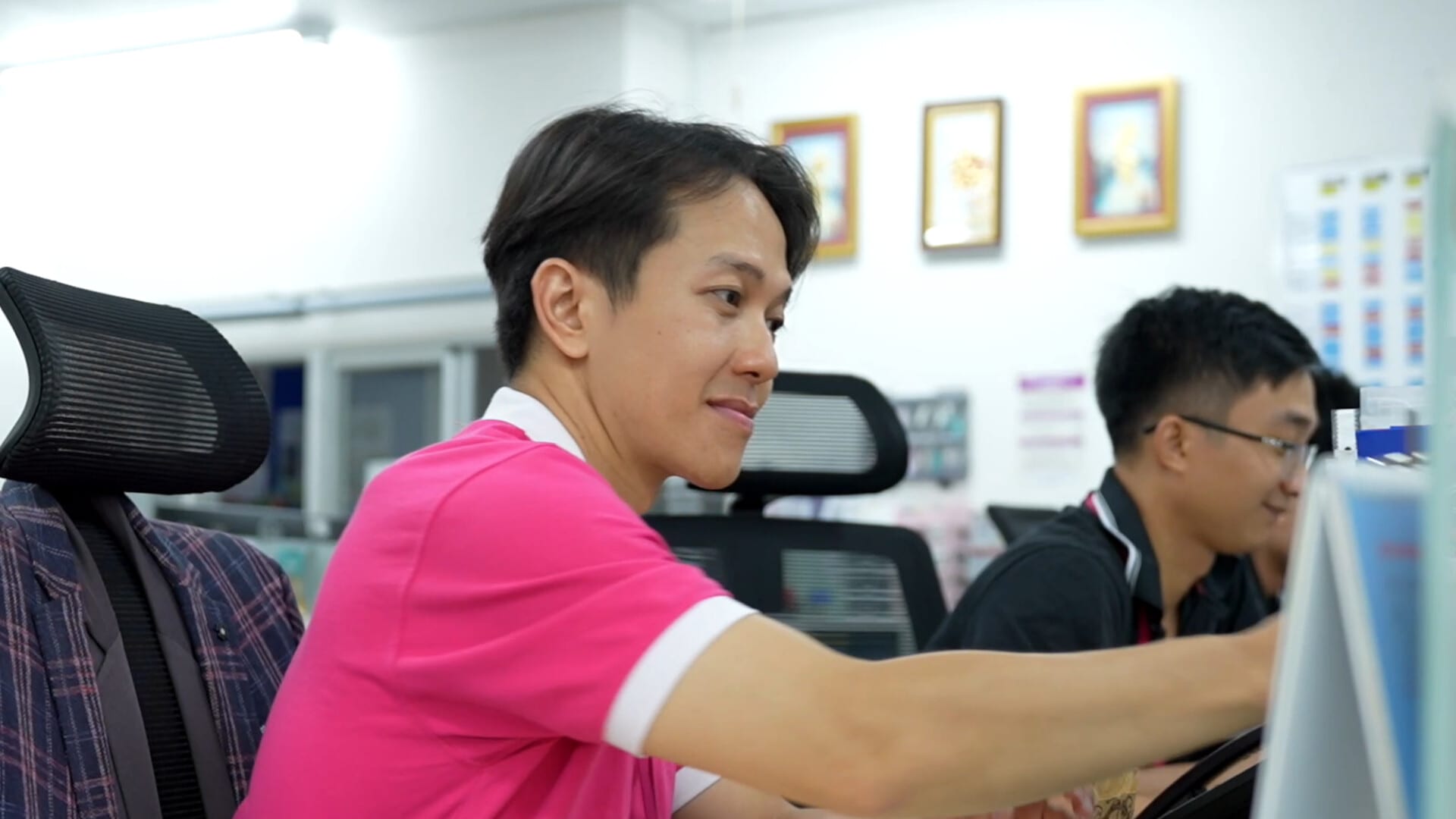 Người đàn ông áo hồng ngồi làm việc bên máy tính, đồng nghiệp làm việc phía sau