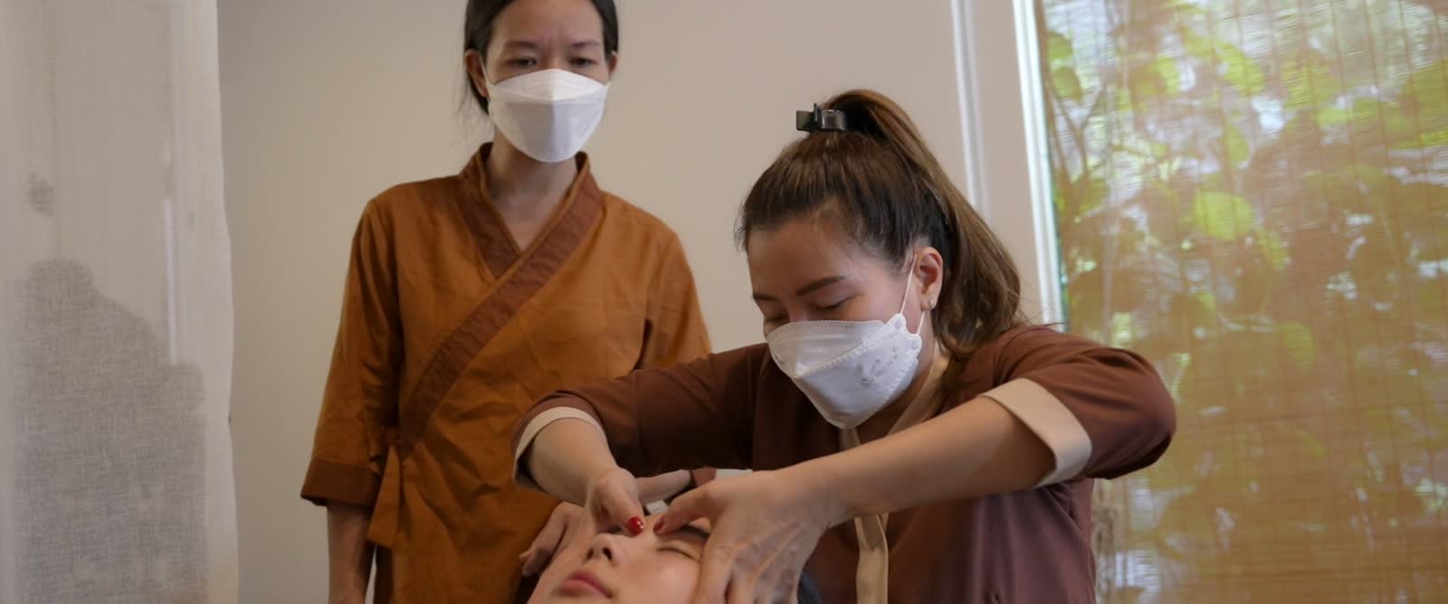 Kỹ thuật viên spa chăm sóc da mặt cho khách, một nhân viên khác đứng quan sát