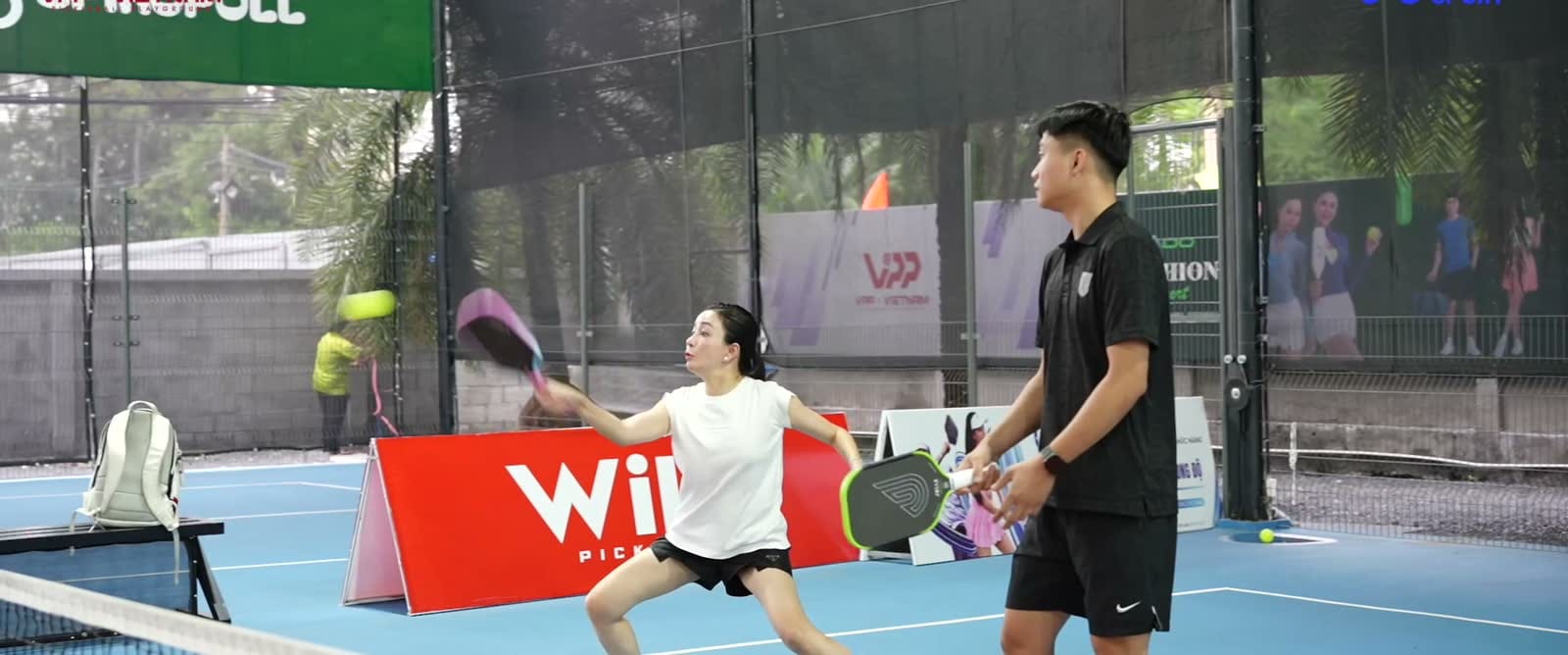 Đôi nam nữ đánh pickleball trên sân có mái, biển quảng cáo ven sân