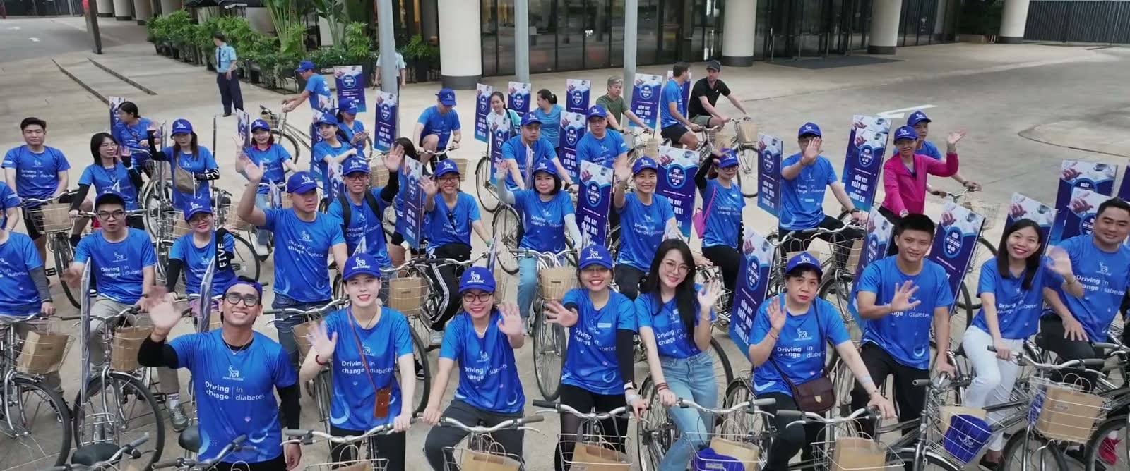 Nhân viên áo xanh ngồi trên xe đạp xếp hàng trước toà nhà, giơ tay chào