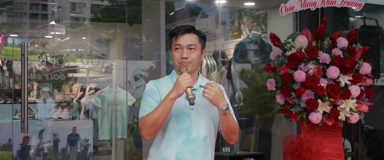 Người phát biểu cầm micro trước cửa hàng ADIDAS Golf và các lẵng hoa chúc mừng.